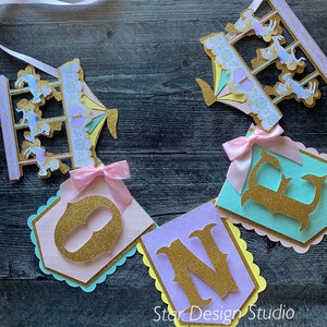 May include: A colorful banner with the word "ONE" in gold glitter letters. The banner is decorated with pastel pink, blue, and yellow paper cutouts of a carousel with horses. The banner is perfect for a first birthday party.