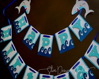 Peut inclure: Une bannière en papier bleue et turquoise avec les mots "Happy Birthday" en lettres blanches. La bannière est décorée de vagues bleues et d'un ruban bleu. Un requin en papier bleu et gris est suspendu au-dessus de la bannière.
