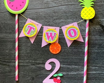 May include: A pink and green cake topper with a banner that says "TWO" and a number two with cherries. The banner is strung between two straws with a watermelon and a pineapple on top. The text "Star Design Studio" is at the bottom of the image.