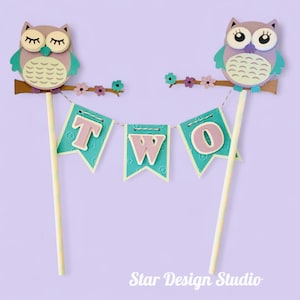May include: Two owl-shaped cake toppers with a banner that spells "TWO". The owls are purple, teal, and beige, perched on brown branches with pink flowers. The banner flags are teal with pink letters. The background is a light purple.
