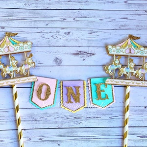 May include: A cake topper set with two gold glitter carousel toppers and a banner that says "ONE" in gold glitter letters. The banner is made of pink, purple, and turquoise paper.
