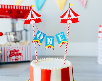 Puede incluir: Un pastel de rayas blancas y rojas con glaseado blanco y un adorno de pastel con temática de circo. El adorno presenta dos carpas de rayas rojas y blancas con banderas amarillas y una pancarta azul que dice "ONE".