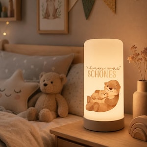 Könnte beinhalten: Eine zylindrische Tischleuchte mit warmem Lichtschein, verziert mit einer Illustration von zwei Ottern und dem Text "Träum was SCHÖNES". Die Leuchte hat einen glatten, weißen, lichtdurchlässigen Schirm und einen schlichten, hellen Sockel.
