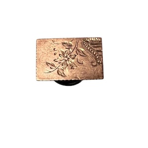 May include: A rectangular copper-toned metal object featuring an engraved floral design on its surface. The piece displays a central flower with leaves and a textured, patterned border along the right side, with a dark circular base visible underneath.