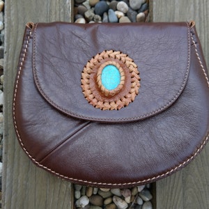 以下が含まれることがあります： 茶色の革製のハンドバッグ。フラップクロージャーと、茶色の革で囲まれた装飾的なターコイズストーンが特徴です。