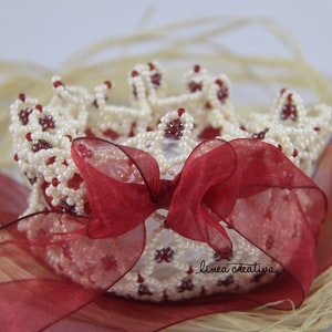 May include: A white beaded crown with red accents, tied with a red ribbon bow. The crown is sitting on a bed of straw.
