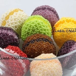 Może przedstawiać: Kolekcja kolorowych, koralikowych makaroników w szklanej misce. Makaroniki są wykonane z małych koralików i występują w odcieniach fioletu, zieleni, żółci, bieli, brązu i różu.