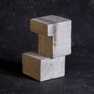 Peut inclure: Deux blocs de béton gris clair empilés. Le bloc supérieur a une découpe rectangulaire, créant une forme géométrique intéressante. Les blocs ont une surface rugueuse et texturée, sur un fond sombre et moucheté.