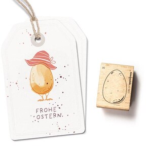 Pode incluir: Um cartão branco com um ovo castanho a usar um chapéu rosa e o texto "Frohe Ostern", que significa Feliz Páscoa em alemão. O cartão está ao lado de um carimbo de madeira com um desenho de ovo castanho.