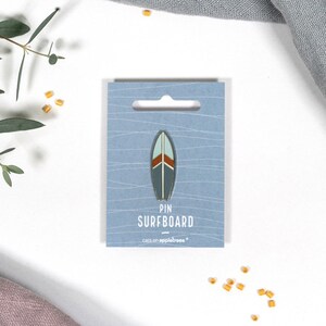 Könnte beinhalten: Eine Surfbrett-Anstecknadel auf einer hellblauen Karte. Die Nadel hat die Form eines Surfbretts mit dunkelblauem Grund, hellblauem Zentrum und braunen und weißen Akzenten. Auf der Karte steht "PIN SURFBOARD" und "cats on appletrees".