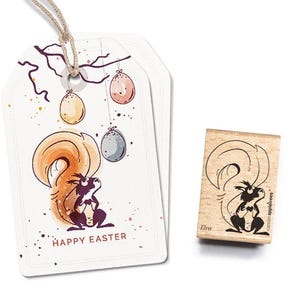Könnte beinhalten: Eine weiße Karte mit einer bunten Illustration eines Eichhörnchens, das ein Osterei hält. Die Karte sagt "Frohe Ostern". Ein Holz-Stempel mit einer schwarzen Silhouette eines Eichhörnchens ist ebenfalls abgebildet. Der Stempel sagt "Ella" und "Craftables & Appliques".