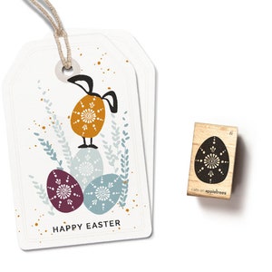 Può includere: Un'etichetta regalo bianca con un coniglio che sta in piedi su tre uova di Pasqua decorate. L'etichetta dice "HAPPY EASTER" e ha accenti dorati e blu. È mostrato anche un piccolo timbro di legno con un design di uovo bianco e nero. Il timbro dice "cats on appletrees" e ha il numero "6" su di esso.