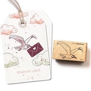 Könnte beinhalten: Ein weißes Etikett mit einer Cartoon-Möwe, die einen burgunderfarbenen Umschlag mit einem Herzen trägt. Das Etikett trägt die Aufschrift "SENDING LOVE" und ist mit Wolken verziert. Ein Holzstempel mit dem gleichen Möwen-Design ist ebenfalls abgebildet.