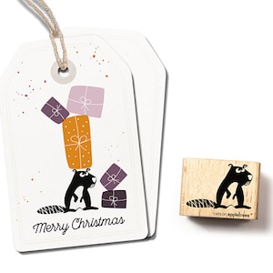 Könnte beinhalten: Ein weißer Geschenkanhänger mit einer Schwarzweißillustration eines Bibers, der Geschenke trägt. Der Text "Merry Christmas" ist auf dem Anhänger gedruckt. Der Anhänger ist an einer Schnur befestigt. Ein Holzstempel mit einer Schwarzweißillustration eines Bibers ist unter dem Anhänger abgebildet. Der Text "cats on appletrees" ist auf dem Stempel gedruckt.