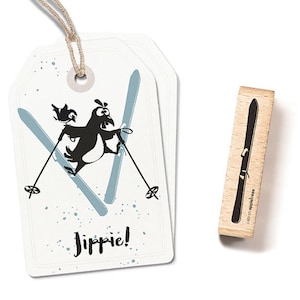 May include: Two white gift tags featuring a cartoon penguin skiing, with a small bird. The tags have a string for hanging and the word "Jippie!" is written on the tag. A wooden ski stamp is also included.