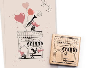 Puede incluir: Un sello de goma de madera con una ilustración en blanco y negro de una pequeña tienda con un toldo a rayas. La tienda tiene un letrero que dice "Einladung" y hay dos pájaros pequeños con notas musicales sobre la tienda. El sello está sobre un fondo marrón claro.
