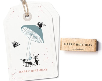 Può includere: Un'etichetta regalo bianca con un fungo blu e formiche e mosche nere. L'etichetta ha pois rosa e il testo "HAPPY BIRTHDAY". È incluso anche un timbro di legno con il testo "HAPPY BIRTHDAY".
