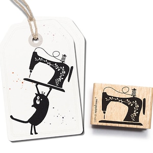 May include: A black cat holding up a vintage sewing machine with a floral design. The image is a rubber stamp with the text "cats on appletrees" on the side.