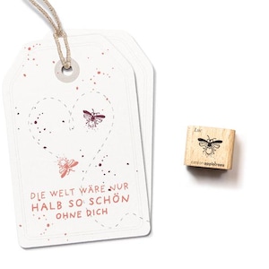 Könnte beinhalten: Ein weißer Geschenkanhänger mit einer Herzform aus gepunkteten Linien und zwei Bienenillustrationen. Der Text auf dem Anhänger lautet "Die Welt wäre nur halb so schön ohne dich". Ein kleiner Holzstempel mit einer Bienenillustration und dem Text "Luc" ist ebenfalls abgebildet.
