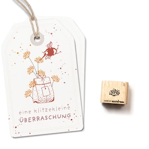 May include: A white gift tag with a brown string attached. The tag has a brown and yellow illustration of a bee flying towards a small gift bag with a brown bow. The text on the tag reads "eine klitzekleine ÜBERRASCHUNG".