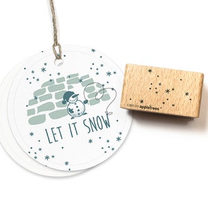 Könnte beinhalten: Ein Holzstempel mit einem Schneemann-Motiv und dem Text "Let it snow". Der Stempel befindet sich auf einem weißen Hintergrund mit einem Kreis aus Schneeflocken.