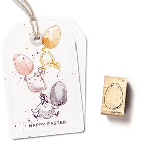Könnte beinhalten: Eine weiße Karte mit einer Aquarell-Illustration von drei Hühnern, die bunte Eier halten. Die Karte lautet "Frohe Ostern" und ist mit einem hellbraunen Rand verziert. Ein kleiner Holz-Stempel mit einem einzelnen Ei-Design ist ebenfalls abgebildet.