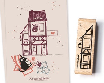 Puede incluir: Ilustración de una casa caprichosa con un gato relajándose en una tumbona, y la frase "La vie est belle!". También se muestra un sello de madera con un diseño de casa. La obra de arte está en tonos marrones y negros.