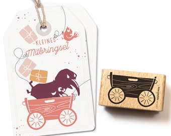 Pode incluir: Um carimbo de madeira com uma silhueta preta de uma carroça carregando presentes. O carimbo é etiquetado como "cats on appletrees". O carimbo vem acompanhado de uma etiqueta de presente com um design semelhante, que apresenta um pássaro e o texto "kleines Mitbringset".