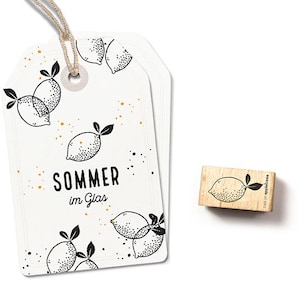 Op de afbeelding: Een wit label met zwarte en gouden citroenillustraties en de tekst "Sommer im Glas". Een houten stempel met een citroenillustratie en de tekst "cats on apple trees" is ook te zien.