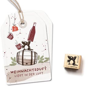 May include: Two white rectangular paper gift tags with a jute string and a small wooden rubber stamp. The tags feature a brown mole standing on a barrel, holding a bottle, with red mugs and the text WEIHNACHTSDUFT LIEGT IN DER LUFT.