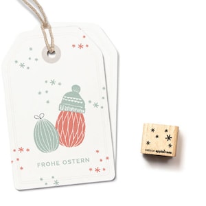 Könnte beinhalten: Ein weißer Geschenkanhänger mit einer roten und grünen Illustration eines Winterhutes und eines Geschenks. Der Text "Frohe Ostern" ist auf dem Anhänger gedruckt. Ein kleiner Holz-Stempel mit dem Text "cats on appletrees" und einem Schneeflocken-Design ist ebenfalls abgebildet.
