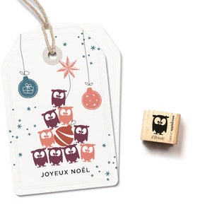 May include: Two white gift tags with a Christmas tree design featuring owls, baubles, and snowflakes. The text "JOYEUX NOËL" is at the bottom. A small wooden stamp with an owl illustration is next to the tags.