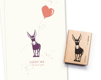 Puede incluir: Una tarjeta de felicitación y un sello de madera con un diseño de burro. La tarjeta muestra un burro morado con un globo en forma de corazón y el texto "LUCKY ME I found you." El sello tiene una ilustración de burro similar.