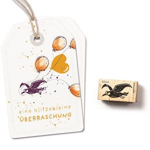 Könnte beinhalten: Zwei weiße Geschenkanhänger mit einer skurrilen Illustration eines Vogels, der mit Luftballons fliegt. Die Anhänger tragen den deutschen Text "eine klitzekleine ÜBERRASCHUNG". Ein Holzstempel mit einer Vogelgrafik und dem Namen "Klaus" ist ebenfalls vorhanden.