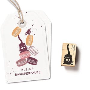 Könnte beinhalten: Zwei weiße Geschenkanhänger mit einer Schnur, die eine Katzenillustration mit Makronen zeigt. Die Anhänger tragen den Text "KLEINE KNUSPERPAUSE". Ein Holzstempel mit einer Katzenillustration und dem Text "Frida cats on appletrees" ist ebenfalls vorhanden.