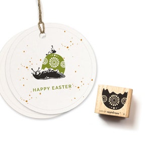 Könnte beinhalten: Ein Holz-Stempel mit einem schwarz-weißen Design einer Schnecke, die ein zerbrochenes Ei mit einem grün-weißen Muster trägt. Der Text "Happy Easter" ist auf einem weißen Etikett mit goldenen Punkten gedruckt. Der Stempel ist mit "crafty appltrees 6" gekennzeichnet.