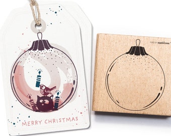 Puede incluir: Un sello de goma de madera con un contorno negro de un adorno navideño con un gato y un pájaro dentro. El adorno está rodeado de un fondo de acuarela en rosa y blanco. El texto "Merry Christmas" está impreso debajo del adorno.