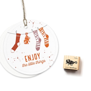 May include: A round white tag with a string attached. The tag has a design of socks hanging on a clothesline with a small fish and the text "ENJOY the little things". A small wooden stamp with a chameleon design and the text "cats on appletrees" is also shown.