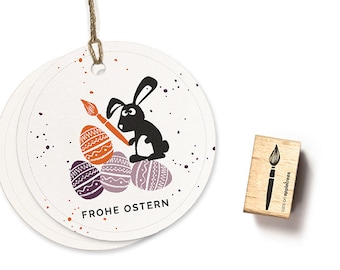 Pode incluir: Uma etiqueta redonda branca com um coelho preto segurando um pincel e pintando ovos de Páscoa laranja e roxo. A etiqueta diz "Frohe Ostern", que é alemão para "Feliz Páscoa".