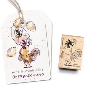 Peut inclure: Une étiquette cadeau blanche avec une illustration dessinée à la main d'un coq tenant un œuf avec le texte "eine klitzekleine ÜBERRASCHUNG". Un tampon en caoutchouc en bois avec une illustration de coq et le texte "cards of appletrees Louis Germain" est également présenté.