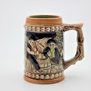 May include: A brown ceramic beer stein with a tree-like handle. The stein features a relief design of two men in traditional clothing drinking from a barrel.