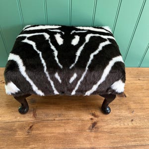 May include: A square footstool featuring a zebra print pattern in black and white. The footstool has dark brown, curved legs. The top is covered in a faux fur material.