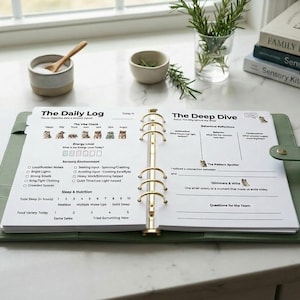 May include: Open green binder with gold rings, filled with daily log pages. The pages have sections for mood, energy levels, and sensory environment. A wooden pen rests on the side. Books and small bowls are in the background.