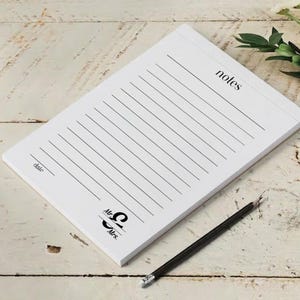 Peut inclure: Bloc-notes blanc avec des pages lignées, le mot "notes" en haut et "date" en bas à gauche. Un crayon noir est posé à côté. Les mots "Mr. & Mrs." sont imprimés en bas. Le bloc-notes est sur une surface en bois clair.