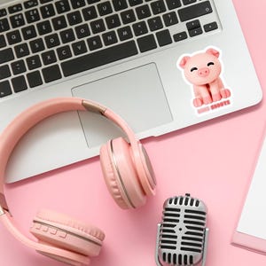 May include: A pink setup featuring a laptop with a pig sticker that reads "BINO SHOOTS", pink headphones, and a silver microphone. The items are arranged on a pink surface, suggesting a workspace or recording setup.