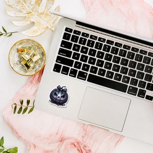 May include: A black and blue Halloween pumpkin sticker with a bow is placed on a silver laptop. The laptop is on a white surface with a pink cloth, a gold leaf, and a small bowl with gold paper clips.