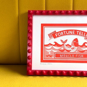 May include: A red framed print with a white background and a red illustration of a fish with the text "FORTUNE TELLER MIRACLE FISH" in white. The print is signed "Miracle Fish" in the bottom left corner.