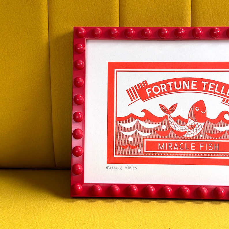 May include: A red framed print with a white background and a red illustration of a fish with the text "FORTUNE TELLER MIRACLE FISH" in white. The print is signed "Miracle Fish" in the bottom left corner.
