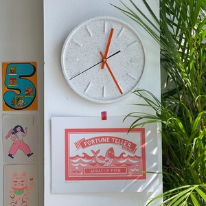 May include: A white clock with black hands and an orange second hand. The clock has a speckled white face and is hanging on a white wall. A red and white print with the text "Fortune Teller Miracle Fish" is hanging on the wall below the clock.
