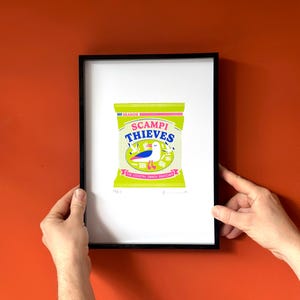 May include: A framed print of a colourful illustration of a bag of crisps called "Scampi Thieves" with a seagull on the front. The bag is green with pink and blue accents. The text on the bag reads "The Coastal Snack Snatchers".